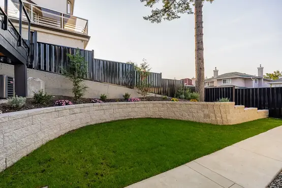 Burnaby South slope -Sloped backyard featuring a concrete retaining wall, Allan block planters, and a black powder-coated metal fence, complemented by lush, well-designed landscaping.