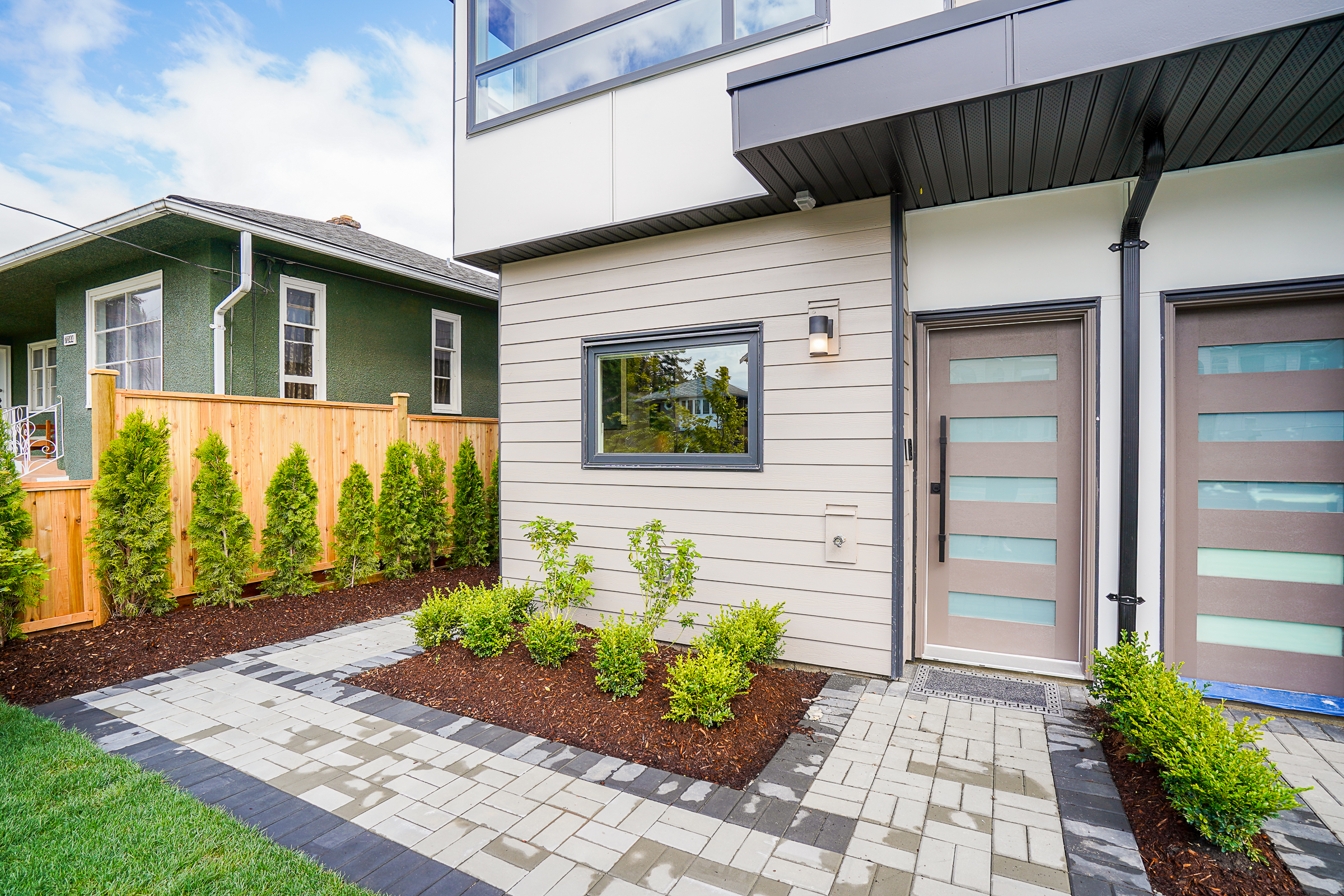 Front yard landscaping at a multiplex featuring a paver pathway, manicured grass lawn, freshly planted trees, and mulched planting beds with flowers and ornamental plants.