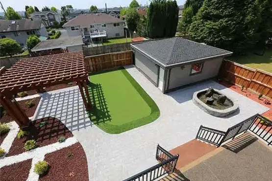 Backyard landscaping in Coquitlam, British Columbia featuring grey concrete pavers, red lava rock accents, a decorative water feature, and a synthetic turf putting green area.