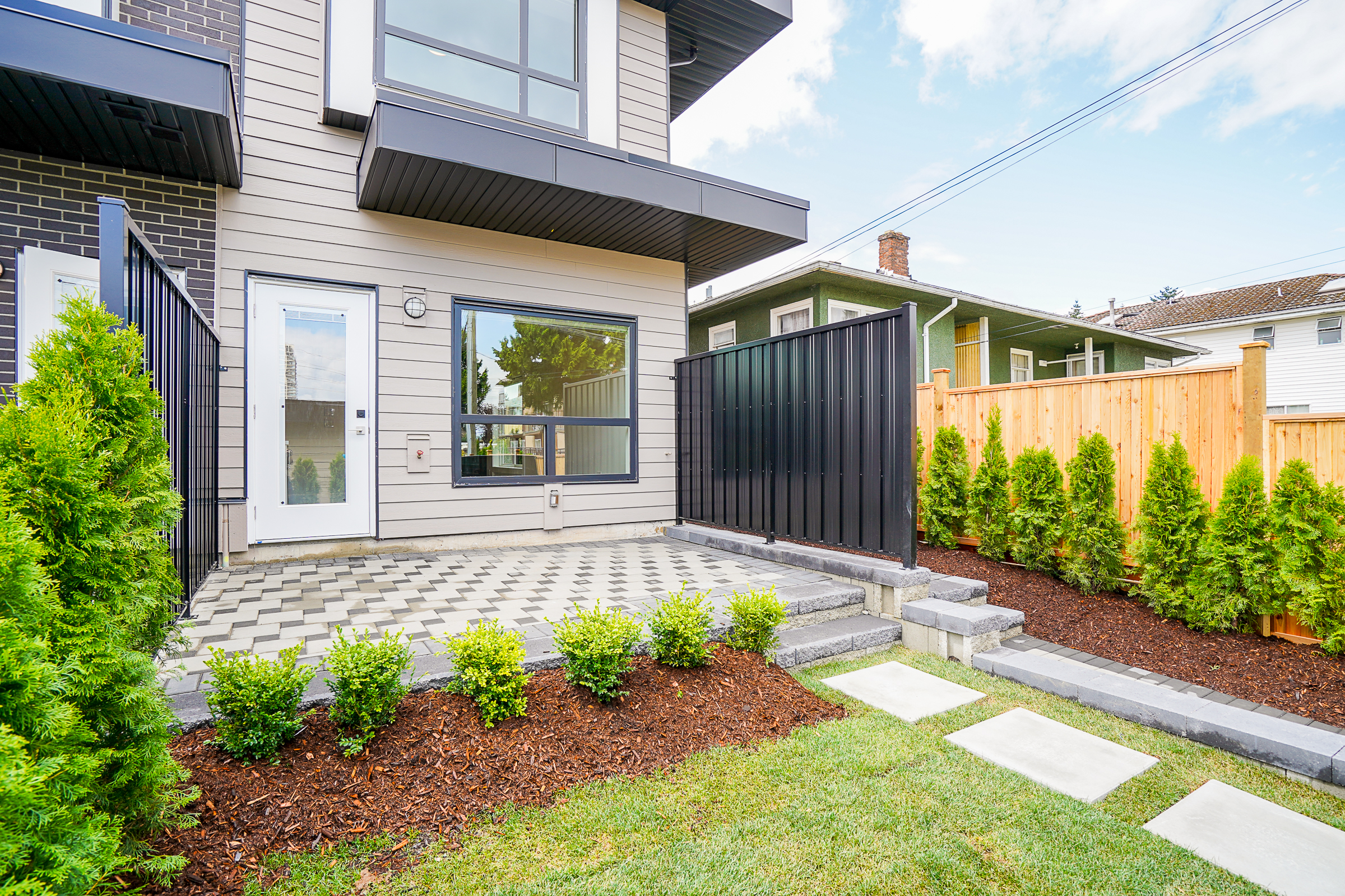Backyard landscaping for a Vancouver multiplex featuring mulched shrub beds, metal and cedar privacy fencing, a concrete step pathway to the garage, and a paver patio landing for a BBQ area.